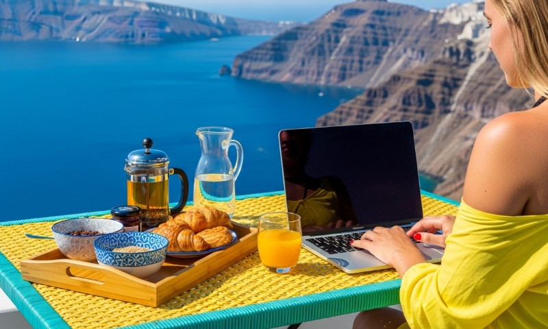 A woman in a yellow top works on a laptop at a table with breakfast